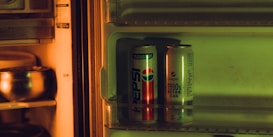 A refrigerator interior lit with a warm, yellow-orange light reveals two retro Pepsi cans on a shelf. The cans feature vintage logos and designs, hinting at a 1990s theme. The faint greenish hue from artificial lighting adds to the nostalgic atmosphere.