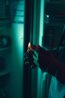 A person is holding a lighter with a small flame in front of an open refrigerator. The lighting is dim and has a greenish tint, giving a moody atmosphere. Shadows are cast on the hands, and various objects are faintly visible inside the refrigerator.