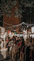 A bustling night market scene with people browsing and interacting with vendors under a canopy of decorative string lights. The market is lined with stalls displaying various goods, including clothing and trinkets, and is shaded by trees and surrounded by a brick building.