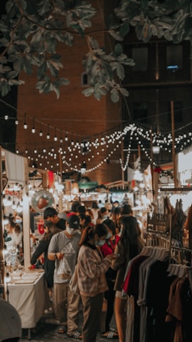 A bustling night market scene with people browsing and interacting with vendors under a canopy of decorative string lights. The market is lined with stalls displaying various goods, including clothing and trinkets, and is shaded by trees and surrounded by a brick building.