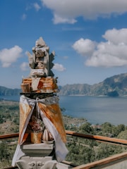 A traditional Balinese shrine adorned with offerings stands prominently in the foreground. It is intricately decorated with a mix of fabrics, baskets, and what appear to be ceremonial items. The background reveals a panoramic view of a placid lake surrounded by verdant hills under a bright blue sky dotted with fluffy white clouds.