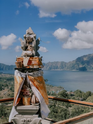 A traditional Balinese shrine adorned with offerings stands prominently in the foreground. It is intricately decorated with a mix of fabrics, baskets, and what appear to be ceremonial items. The background reveals a panoramic view of a placid lake surrounded by verdant hills under a bright blue sky dotted with fluffy white clouds.