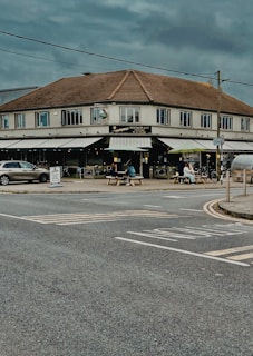 A corner restaurant with outdoor seating is situated at an intersection. The building has a brown sloped roof and large windows. Several people are seated at picnic tables under a shaded area. A few parked cars are visible, and the sky is overcast, creating a moody atmosphere.