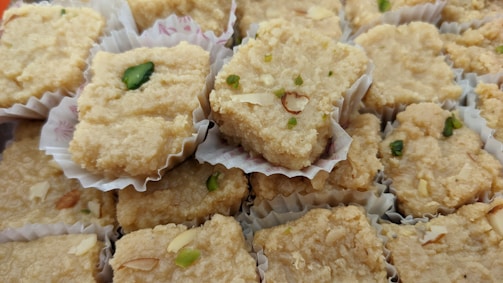 A collection of beige, textured squares resembling pieces of traditional Indian sweets, placed in individual paper cups. Some pieces are garnished with green pistachios and almond slices.