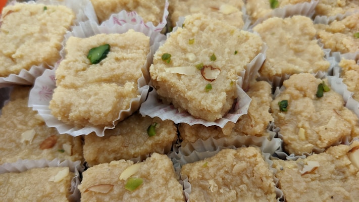A collection of beige, textured squares resembling pieces of traditional Indian sweets, placed in individual paper cups. Some pieces are garnished with green pistachios and almond slices.