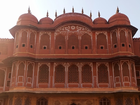 The image features a traditional architectural structure with intricate lattice windows and decorative elements. The building is characterized by its red sandstone facade and symmetrical design, with multiple domes topped with finials. The craftsmanship showcases detailed patterns and motifs typical of historical or culturally significant architecture.