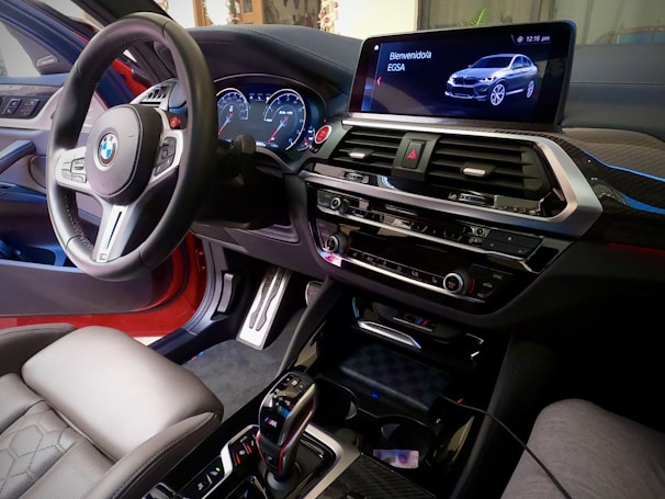 The interior of a BMW car, focusing on the steering wheel, dashboard, and center console. A digital display shows the time and a car image with a message in Spanish. The steering wheel features the BMW logo, and there are various control buttons. The upholstery appears to be leather, and the overall environment is sleek, modern, and high-tech.