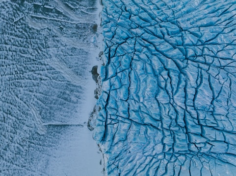 An aerial view of a large glacier with deep cracks and crevices visible in the ice. The image captures the intricate patterns and textures of the blue ice contrasting against the snowy and rocky landscape on the left.