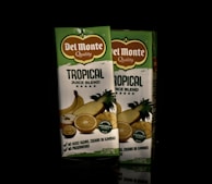 Two cartons of Del Monte Tropical Juice Blend are positioned side by side. The packaging is primarily green and white with images of tropical fruits such as pineapple, orange, and banana. The label highlights the absence of added sugars, colors, flavors, and preservatives.