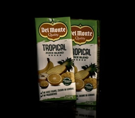 Two cartons of Del Monte Tropical Juice Blend are positioned side by side. The packaging is primarily green and white with images of tropical fruits such as pineapple, orange, and banana. The label highlights the absence of added sugars, colors, flavors, and preservatives.