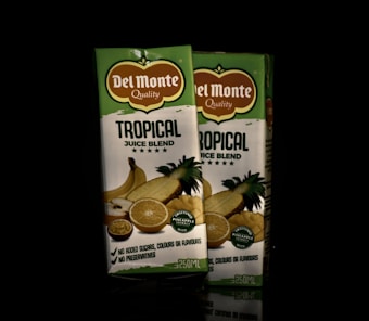 Two cartons of Del Monte Tropical Juice Blend are positioned side by side. The packaging is primarily green and white with images of tropical fruits such as pineapple, orange, and banana. The label highlights the absence of added sugars, colors, flavors, and preservatives.
