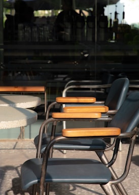 Multiple chairs with metal frames and wooden armrests are aligned beside a circular table. The chairs are positioned on a terrazzo floor with a backdrop of a glass wall, through which a blurred interior space with various objects can be seen.
