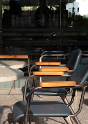 Multiple chairs with metal frames and wooden armrests are aligned beside a circular table. The chairs are positioned on a terrazzo floor with a backdrop of a glass wall, through which a blurred interior space with various objects can be seen.