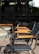 Multiple chairs with metal frames and wooden armrests are aligned beside a circular table. The chairs are positioned on a terrazzo floor with a backdrop of a glass wall, through which a blurred interior space with various objects can be seen.