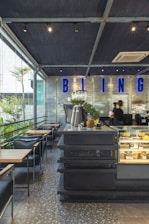 A modern café interior with a sleek, industrial design. The space includes a counter with a black perforated metal surface, a glass display case with pastries, and plants for decoration. Blue neon lettering with the word 'BLING' adorns a silver metallic wall. Several tables and chairs with dark upholstery are arranged on a terrazzo floor. A large window provides natural light and a view of greenery outside.