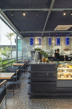 A modern café interior with a sleek, industrial design. The space includes a counter with a black perforated metal surface, a glass display case with pastries, and plants for decoration. Blue neon lettering with the word 'BLING' adorns a silver metallic wall. Several tables and chairs with dark upholstery are arranged on a terrazzo floor. A large window provides natural light and a view of greenery outside.