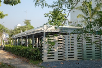 A modern building structure with an open, airy design surrounded by lush green vegetation and trees. The building features a combination of concrete and metal with specific architectural elements such as slatted panels. The scene is complemented by a clear sky, suggesting a pleasant day.