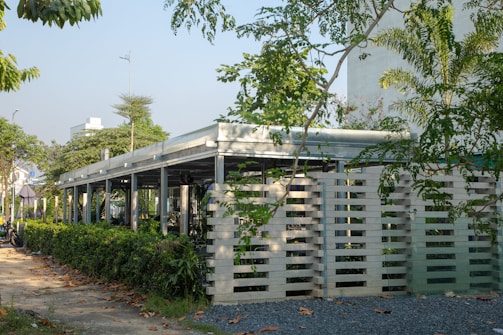 A modern building structure with an open, airy design surrounded by lush green vegetation and trees. The building features a combination of concrete and metal with specific architectural elements such as slatted panels. The scene is complemented by a clear sky, suggesting a pleasant day.