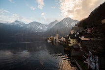 A picturesque lakeside village is nestled among snow-capped mountains, with quaint buildings lining the shore. A prominent church steeple rises in the center, surrounded by traditional alpine houses. The tranquil water reflects the sky and the landscape, while soft clouds capture the warm hues of the setting or rising sun.