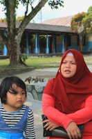 A woman wearing a red hijab and a red outfit is sitting beside a young girl with short black hair wearing a blue dress with black and white striped sleeves. They are outdoors, with a background of trees and a building with blue columns and a tiled roof.