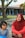 A woman wearing a red hijab and a red outfit is sitting beside a young girl with short black hair wearing a blue dress with black and white striped sleeves. They are outdoors, with a background of trees and a building with blue columns and a tiled roof.