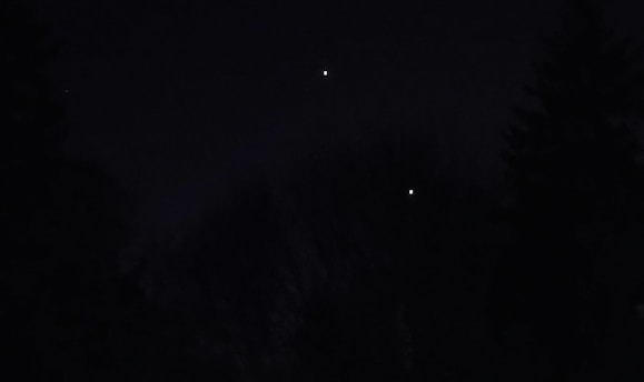 A dark night sky with two bright objects resembling stars or planets. Silhouettes of trees are barely visible against the dark background.