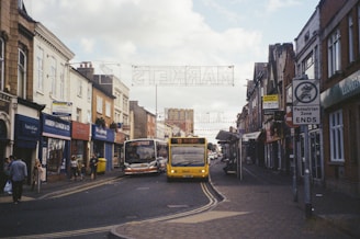 A busy urban street scene with shops lining both sides. Several buses are traveling down the road, and pedestrians are walking along the sidewalks. The architecture is a mix of traditional and modern, with signs indicating a pedestrian zone and a market nearby.