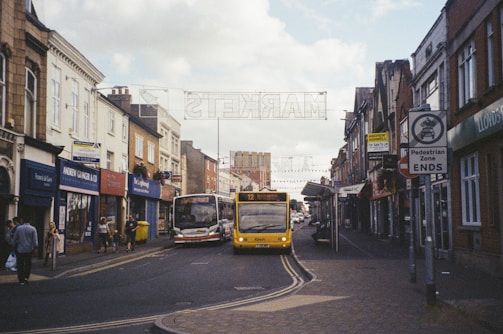 A busy urban street scene with shops lining both sides. Several buses are traveling down the road, and pedestrians are walking along the sidewalks. The architecture is a mix of traditional and modern, with signs indicating a pedestrian zone and a market nearby.
