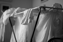 Clothes hang on a drying rack, with a focus on a shirt draped over the metal frame. The monochromatic setting highlights the texture of the fabric and the structure of the drying rack.