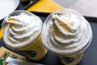 Two cold beverages with creamy whipped topping are placed in plastic cups with dome lids, set on a dark serving tray with a wooden table underneath.