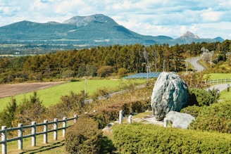A scenic landscape featuring a large rock in the foreground surrounded by greenery, with pathways and a wooden fence. Solar panels are partially visible behind the foliage. A winding road runs through the lush woodland, leading towards distant mountains under a sky filled with scattered clouds.