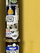 A vertical metal pole is covered with a variety of colorful stickers against a yellow wall. The stickers include text, images, and designs, such as a character wearing an orange jacket and a blue helmet, promotional branding, and phrases like 'HELLO MY NAME IS FROSTY' and 'The SPANTS'. The overall appearance suggests a blend of artistic expression and urban street culture.