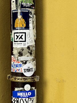 A vertical metal pole is covered with a variety of colorful stickers against a yellow wall. The stickers include text, images, and designs, such as a character wearing an orange jacket and a blue helmet, promotional branding, and phrases like 'HELLO MY NAME IS FROSTY' and 'The SPANTS'. The overall appearance suggests a blend of artistic expression and urban street culture.