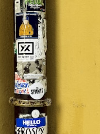 A vertical metal pole is covered with a variety of colorful stickers against a yellow wall. The stickers include text, images, and designs, such as a character wearing an orange jacket and a blue helmet, promotional branding, and phrases like 'HELLO MY NAME IS FROSTY' and 'The SPANTS'. The overall appearance suggests a blend of artistic expression and urban street culture.