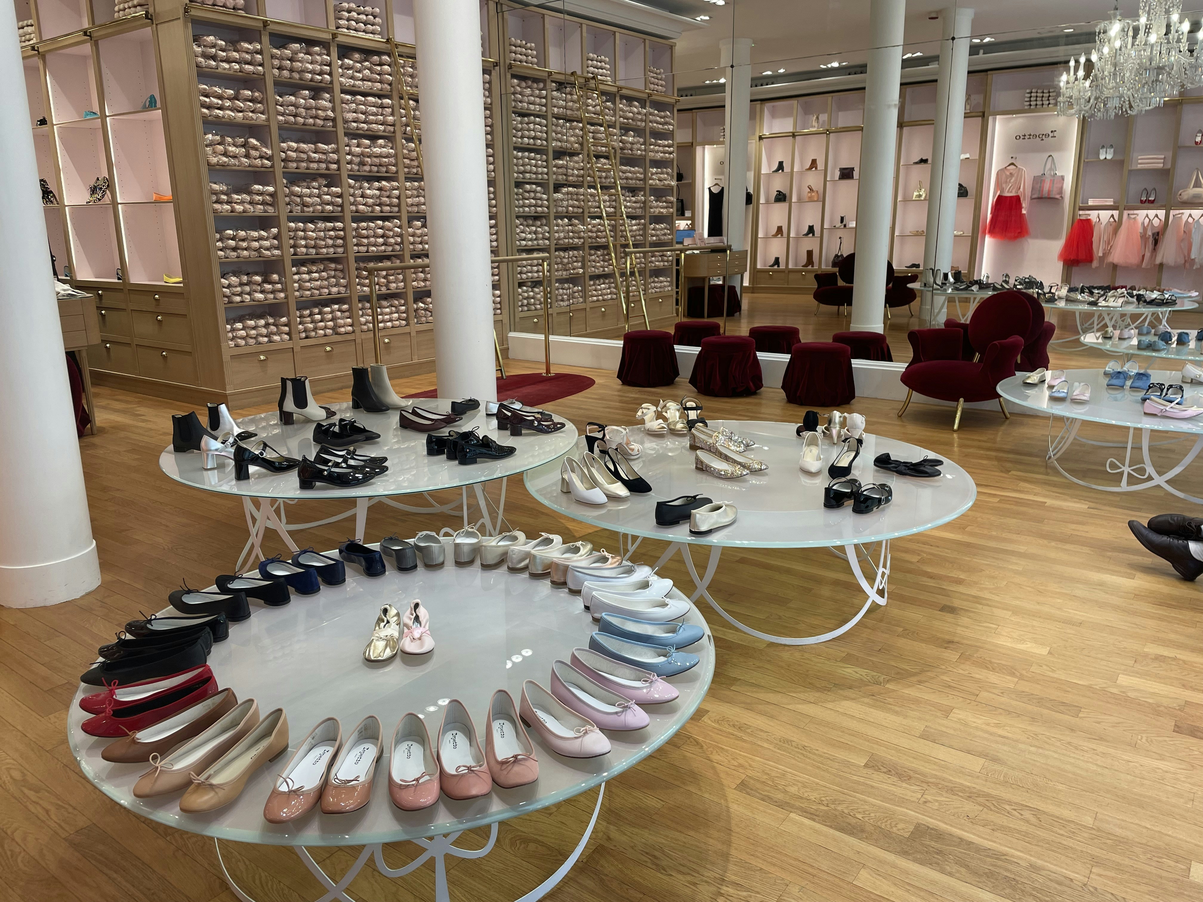 A chic showroom with stylish furniture and elegant displays of women's footwear, featuring flats, sandals, and boots. The wooden flooring and luxurious red chairs create a sophisticated ambiance, with a chandelier adding a touch of elegance. Shelves in the background appear to store stacked ballet shoes.