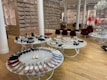 A chic showroom with stylish furniture and elegant displays of women's footwear, featuring flats, sandals, and boots. The wooden flooring and luxurious red chairs create a sophisticated ambiance, with a chandelier adding a touch of elegance. Shelves in the background appear to store stacked ballet shoes.