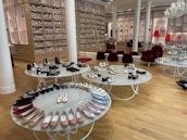 A chic showroom with stylish furniture and elegant displays of women's footwear, featuring flats, sandals, and boots. The wooden flooring and luxurious red chairs create a sophisticated ambiance, with a chandelier adding a touch of elegance. Shelves in the background appear to store stacked ballet shoes.
