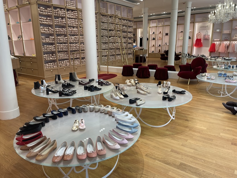 A chic showroom with stylish furniture and elegant displays of women's footwear, featuring flats, sandals, and boots. The wooden flooring and luxurious red chairs create a sophisticated ambiance, with a chandelier adding a touch of elegance. Shelves in the background appear to store stacked ballet shoes.