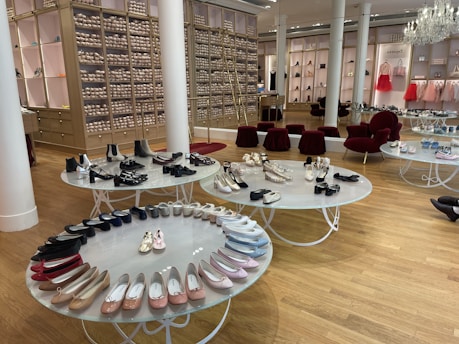 A chic showroom with stylish furniture and elegant displays of women's footwear, featuring flats, sandals, and boots. The wooden flooring and luxurious red chairs create a sophisticated ambiance, with a chandelier adding a touch of elegance. Shelves in the background appear to store stacked ballet shoes.