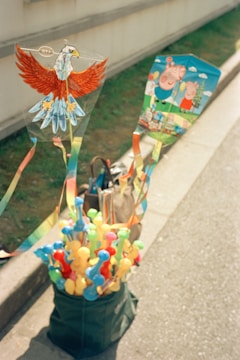 Two kites are displayed with one designed as an eagle and the other featuring cartoon characters. They are attached to a stand filled with colorful toys or balloons. The scene is set outdoors on a sunny day, likely on a pavement near a grassy area. A bag is also placed beside the stand.
