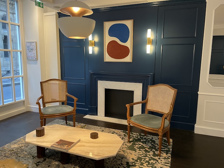 A stylish, modern interior featuring a pair of wooden chairs with cane seats flanking a fireplace. Above the fireplace hangs an abstract artwork in blue and red tones. The room has deep blue-paneled walls with contemporary light fixtures, a marble-topped coffee table, and a patterned rug. Large windows on one side let in natural light.