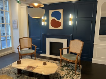 A stylish, modern interior featuring a pair of wooden chairs with cane seats flanking a fireplace. Above the fireplace hangs an abstract artwork in blue and red tones. The room has deep blue-paneled walls with contemporary light fixtures, a marble-topped coffee table, and a patterned rug. Large windows on one side let in natural light.