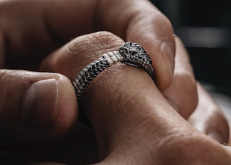 A close-up of hands holding a silver snake ring with intricate scales and a detailed head. The focus is on the craftsmanship of the jewelry against the texture of the skin.