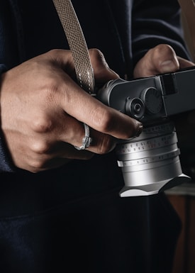 A close-up of hands holding a vintage-style camera with a manual focus lens. The person is wearing a dark-colored clothing and a ring on one finger. The camera has a textured strap wrapped around the hand.