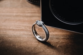 A close-up of a silver ring with a textured snake design placed on a wooden surface. The ring is positioned next to a camera lens with inscriptions visible on its edge, creating a contrast between the metallic textures and the rustic wood.