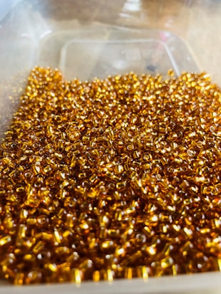 A large quantity of small, shiny, golden beads are clustered together in a plastic container. The beads reflect light, creating a sparkling effect. The container is transparent, and the beads fill it partially.