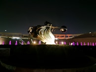 A large bronze-colored sculpture featuring dynamic, flowing lines with an abstract shape is illuminated against a night sky. Surrounding the sculpture are multiple fountains emitting purple light, creating a visually striking contrast. The structure is set within a landscaped area with bushes and hedges, with a backdrop of a partially lit terrace or colonnade.