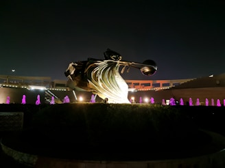 A large bronze-colored sculpture featuring dynamic, flowing lines with an abstract shape is illuminated against a night sky. Surrounding the sculpture are multiple fountains emitting purple light, creating a visually striking contrast. The structure is set within a landscaped area with bushes and hedges, with a backdrop of a partially lit terrace or colonnade.