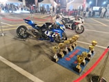 Two high-performance motorcycles are displayed on a parking lot surrounded by red rope barriers. In front of the blue motorcycle, several golden trophies are neatly arranged on a blue mat branded with Suzuki logos. The background features tents, people, and additional motorcycles, suggesting a competitive or exhibition event.