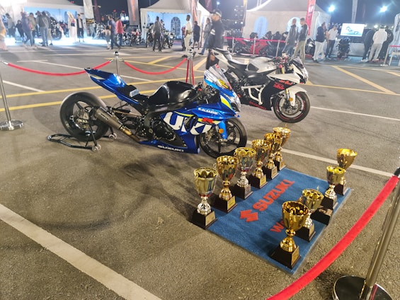 Two high-performance motorcycles are displayed on a parking lot surrounded by red rope barriers. In front of the blue motorcycle, several golden trophies are neatly arranged on a blue mat branded with Suzuki logos. The background features tents, people, and additional motorcycles, suggesting a competitive or exhibition event.